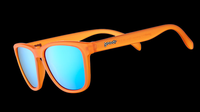 Vista en ángulo de tres cuartos de unas gafas de sol translúcidas de color naranja brillante con cristales reflectantes azules sobre fondo blanco.
