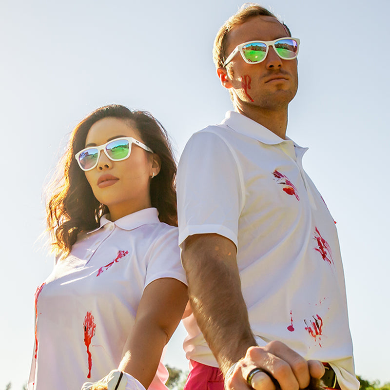 Un homme et une femme portant des lunettes de soleil blanches avec des verres roses se tiennent dos à dos, le sang éclaboussant leurs chemises de golf.