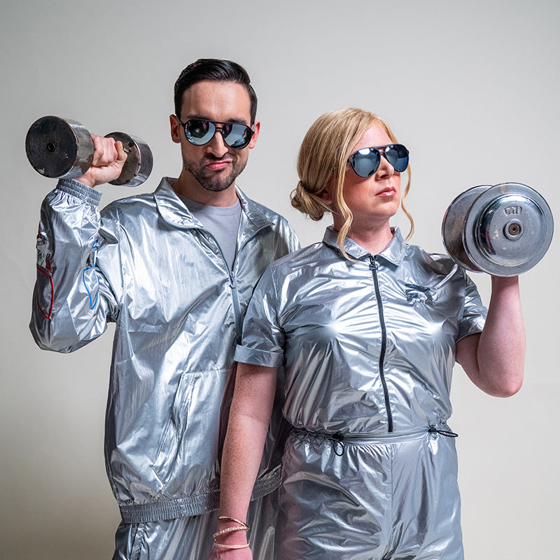 Een man en vrouw met zwarte en chromen pilotenzonnebrillen en zilveren jumpsuits houden barbells vast met felle gezichtsuitdrukkingen.