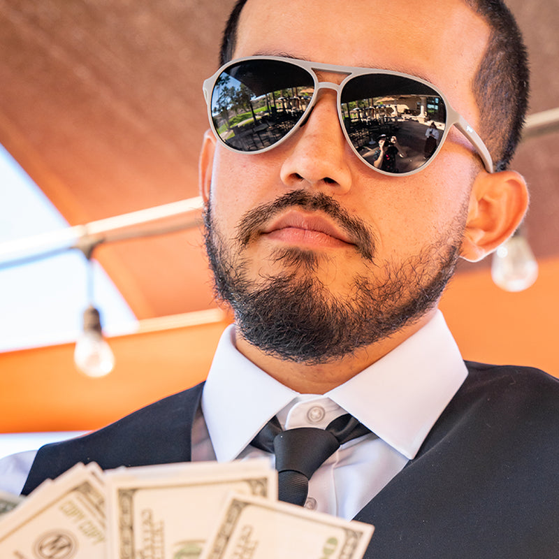 Een man met stropdas en gilet in een golfclubhuis draagt een grijze pilotenzonnebril met zwarte glazen en wappert zichzelf met geld.