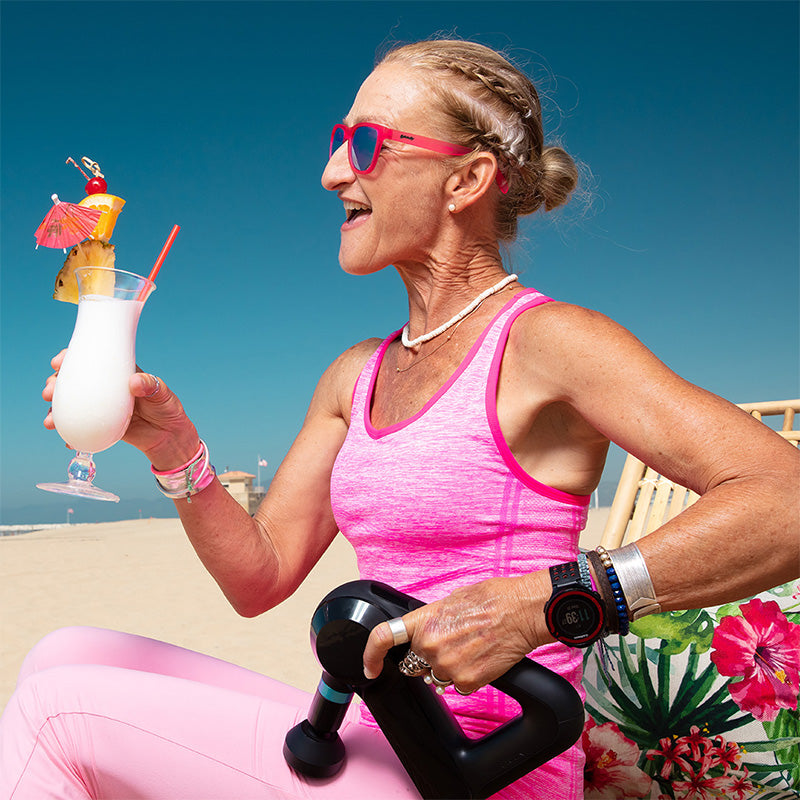Een vrouwelijke hardloopster met een knalroze zonnebril met groene glazen loungt op een strand met een piña colada en een massagepistool voor haar benen.