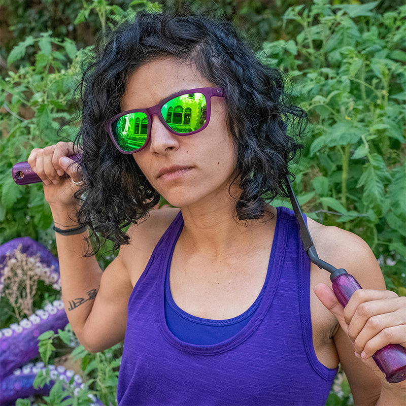 Una donna che indossa occhiali da sole viola con lenti verdi impugna ferocemente attrezzi da giardinaggio, con un tentacolo di polpo alle spalle.