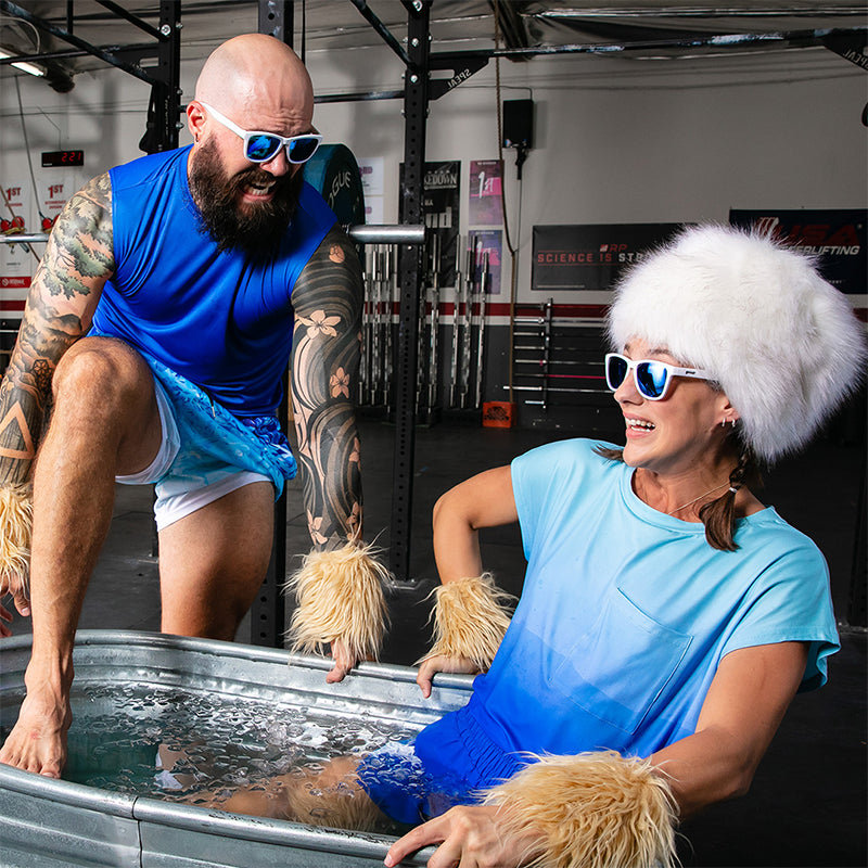 Un homme tatoué s’installe dans un bain de glace à côté d’une femme coiffée d’un chapeau de fourrure blanche, toutes deux portant des lunettes de soleil blanches avec des verres bleus.