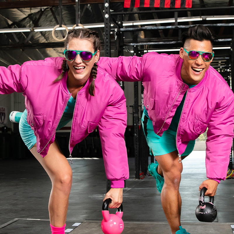Een lachende man en vrouw in roze bomberjacks en groenblauwe vliegeniers met roze reflecterende lenzen doen een kettlebell-training.
