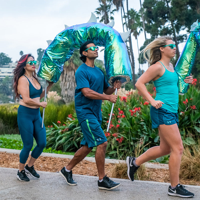 Drei Läufer, die blaugrüne Sonnenbrillen mit blaugrünen Gläsern tragen, halten Teile einer riesigen Loch-Ness-Monsterpuppe, während sie joggen.