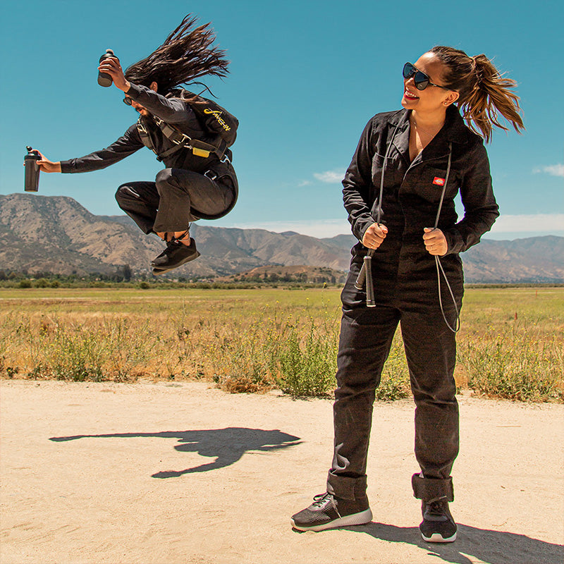 Een parachutist met dubbele eiwitshakes landt in een veld naast een zwarte vrouw in jumpsuit, beiden dragen zwarte vliegenierszonnebrillen.