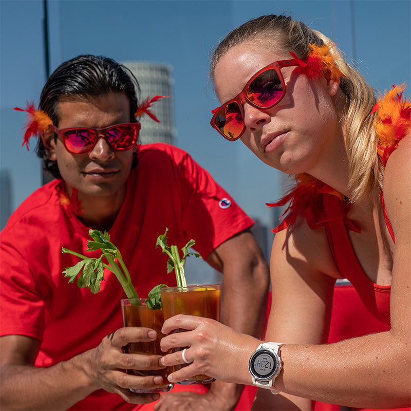 Een man en vrouw met oranje en rode veren en een rode zonnebril met rode reflecterende glazen houden bloody marys vast op een dak.