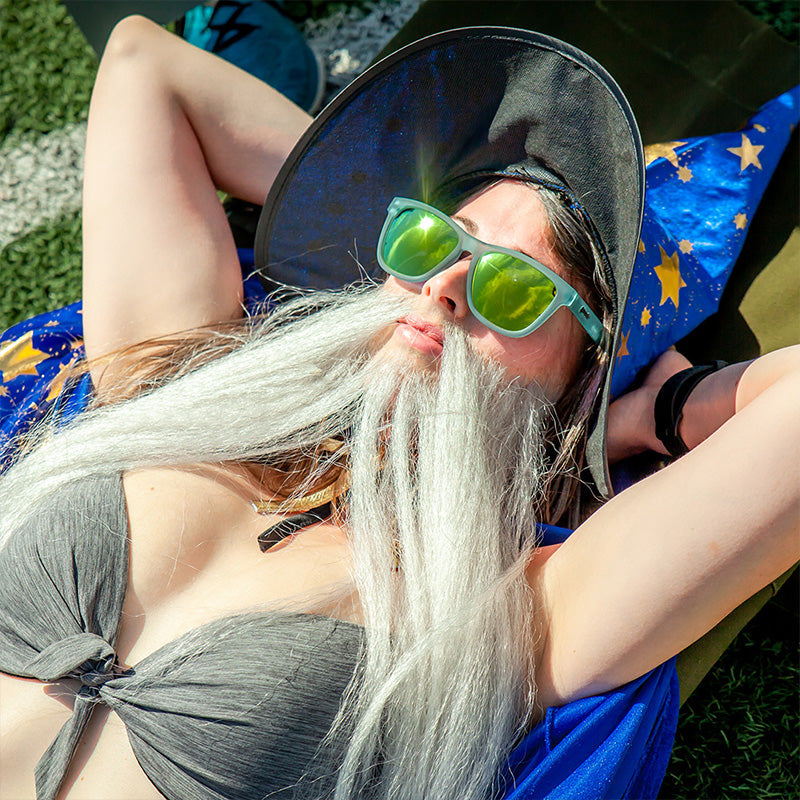 Een vrouw in bikini met een tovenaarshoed & nepbaard zonnebaadt op een atletiekbaan met een lichtblauwe zonnebril met gouden glazen.