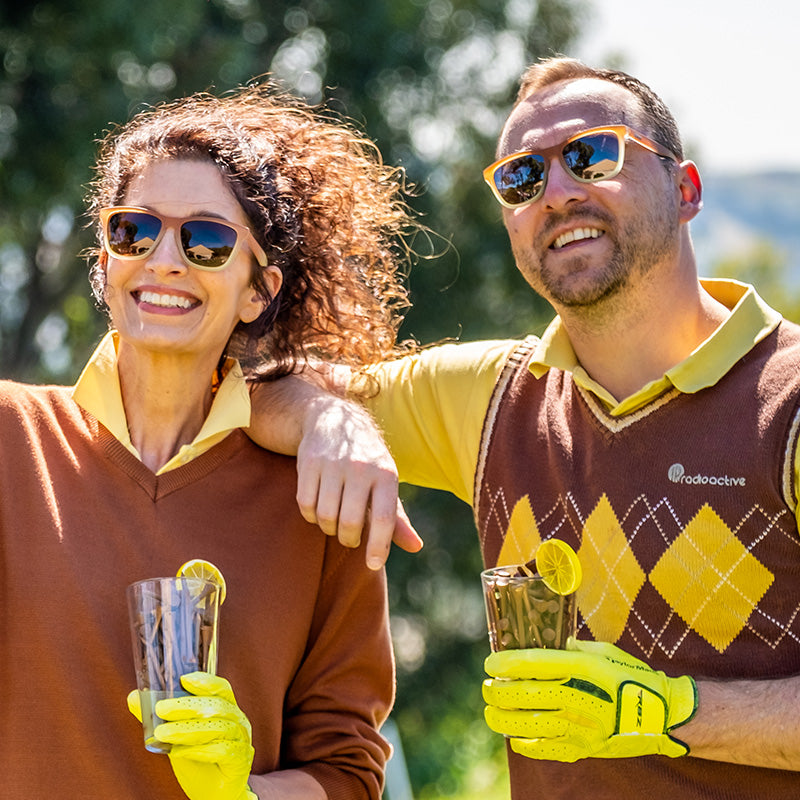 Ein Mann und eine Frau, die eine Sonnenbrille mit braunem Farbverlauf und braunen Gläsern tragen, lächeln auf einem Golfplatz und halten Eistee.