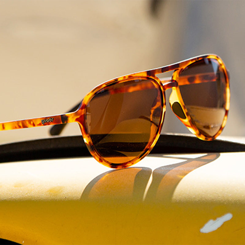 Vista di tre quarti di occhiali da sole da aviatore in tartaruga marrone con lenti marroni non riflettenti appoggiati su una superficie metallica gialla.