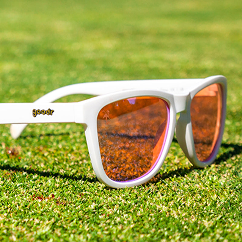 Vista en ángulo de tres cuartos de unas gafas de sol blancas con cristales tintados de rosa sobre un green de golf manchado de sangre.