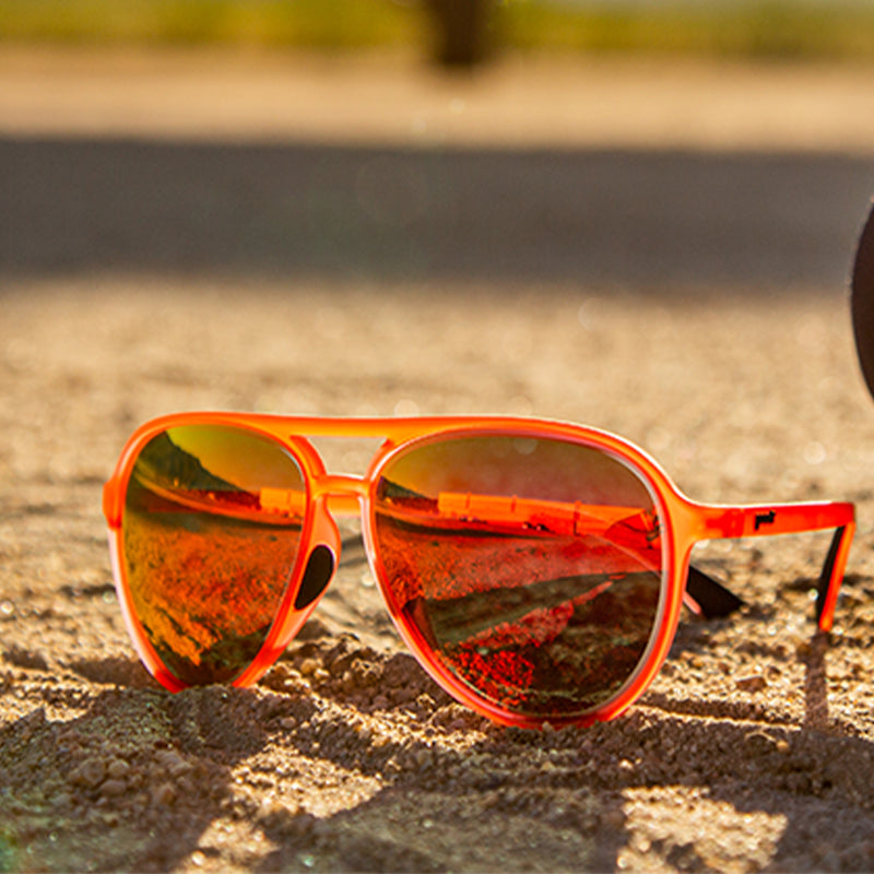 Vista di tre quarti di occhiali da sole rossi da aviatore con lenti riflettenti rosso vivo, posati sulla sabbia lungo la pista di un aeroporto.