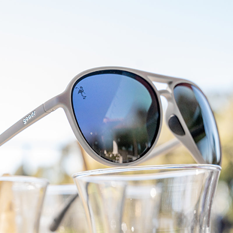 Vista di tre quarti di occhiali da sole da aviatore grigi con lenti nere, appollaiati in cima a un espositore di occhiali da cocktail trasparenti.