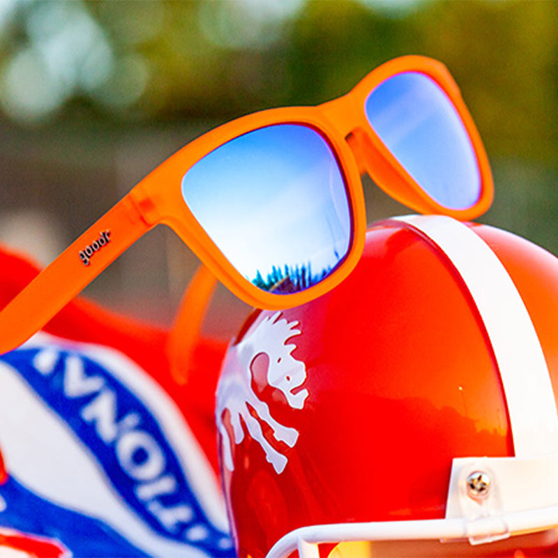 Dreiviertelansicht einer leuchtend orangefarbenen Sonnenbrille mit blauen reflektierenden Gläsern, die auf einem leuchtend orangefarbenen Fußballhelm sitzt.