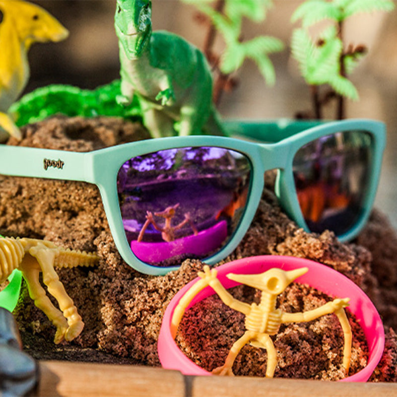 Vue de trois quarts d'angle de lunettes de soleil bleues avec des verres réfléchissants violets, posées sur un monticule de terre à côté de jouets de dinosaures.