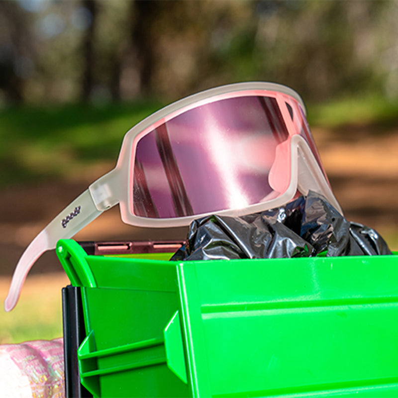 Vue de trois quarts d'angle de lunettes de soleil enveloppantes transparentes avec un seul verre teinté en rose, posées sur une minuscule benne à ordures.