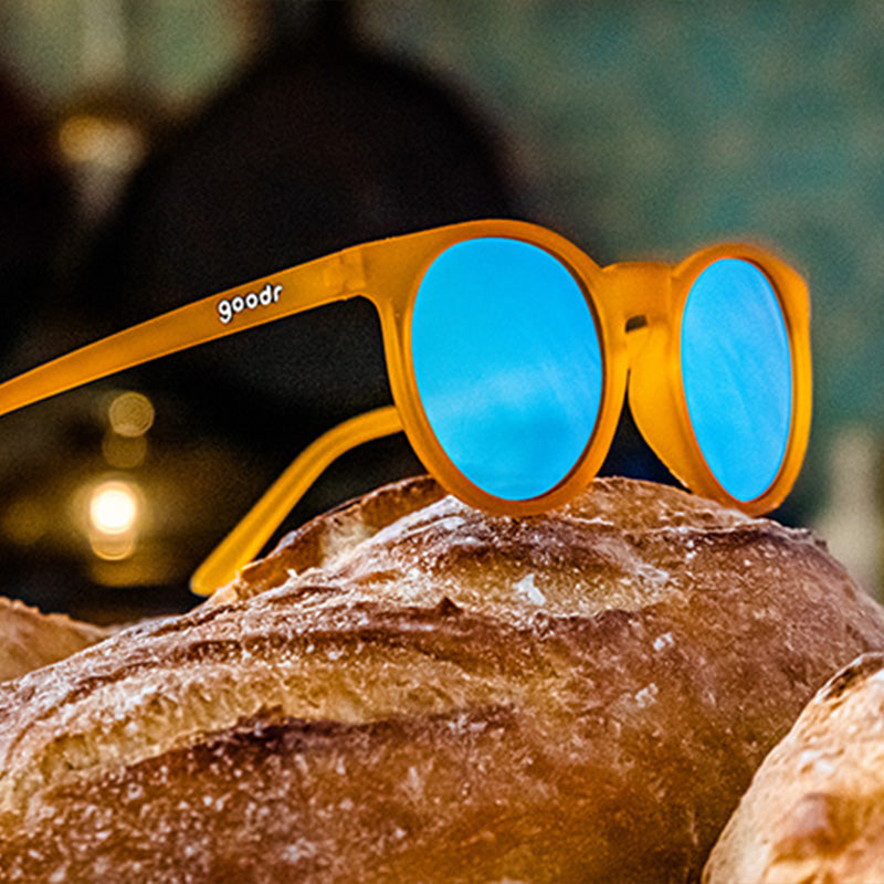 Driekwart bovenaanzicht van een ronde oranje zonnebril met lichtblauwe reflecterende glazen bovenop een versgebakken brood.