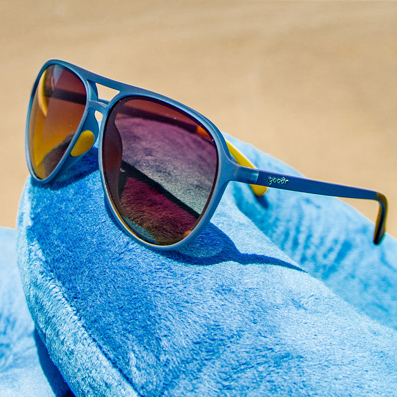 Vista en ángulo de tres cuartos de unas gafas de sol de aviador azul oscuro con cristales ámbar oscuro que descansan sobre una almohada de cuello de viaje de peluche azul.