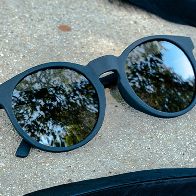 Vista di tre quarti di occhiali da sole rotondi neri con lenti nere non riflettenti, appoggiati sul cemento.