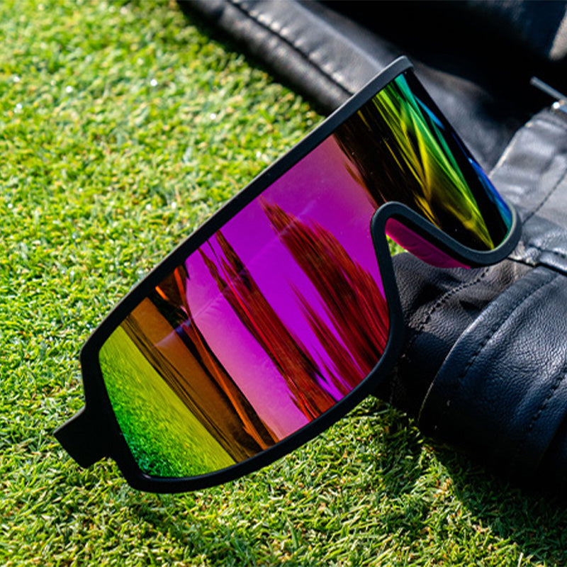 Dreiviertelansicht einer schwarzen Wraparound-Sonnenbrille mit rosafarbenen, reflektierenden Gläsern, die auf einer Lederjacke auf dem Rasen sitzt.