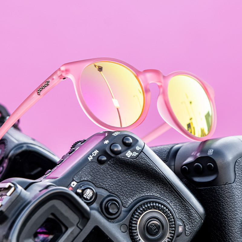 Vista di tre quarti di occhiali da sole rotondi di colore rosa con lenti riflettenti rosa, seduti in cima a una pila di macchine fotografiche.