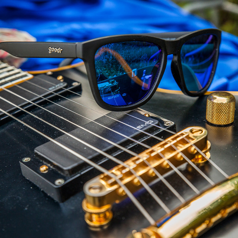 Vista di tre quarti di occhiali da sole neri di forma quadrata con lenti blu riflettenti seduti in cima a una chitarra elettrica.