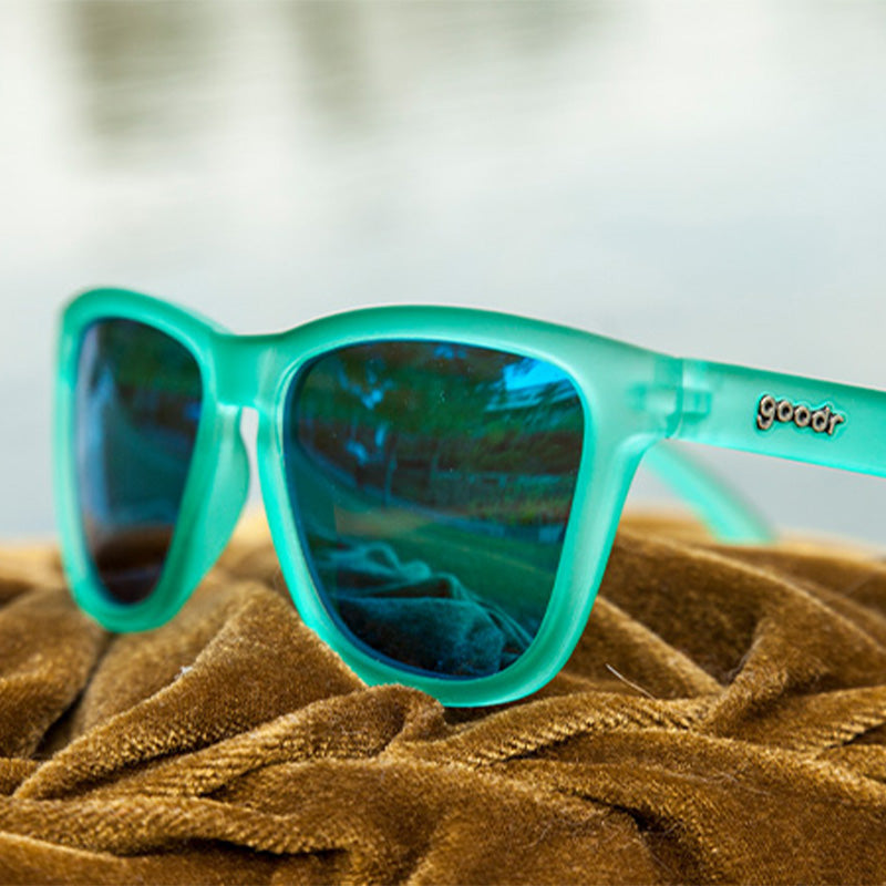 Driekwartaanzicht van een vierkante groenblauwe zonnebril met groenblauwe reflecterende glazen bovenop een stapel goudfluwelen stof.