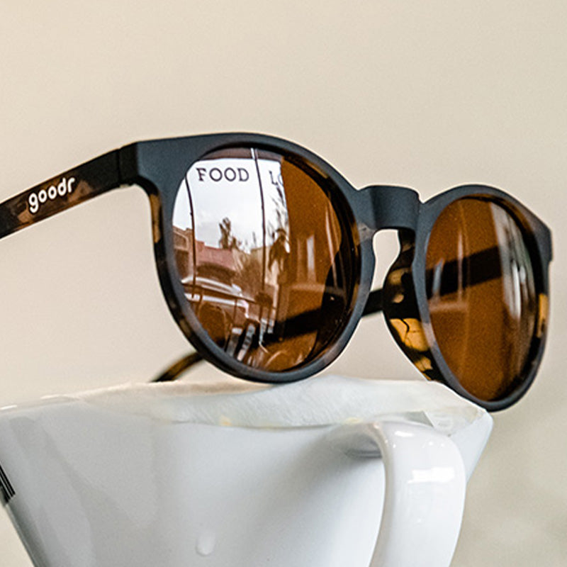 Vue de trois quarts d'angle de lunettes de soleil rondes brunes avec des verres non réfléchissants bruns, posées sur une cafetière à bec verseur.