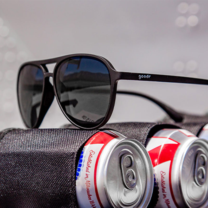 Vista en ángulo de tres cuartos de unas gafas de sol de aviador negras con lentes negras no reflectantes colocadas sobre latas de cerveza en una funda multiusos.