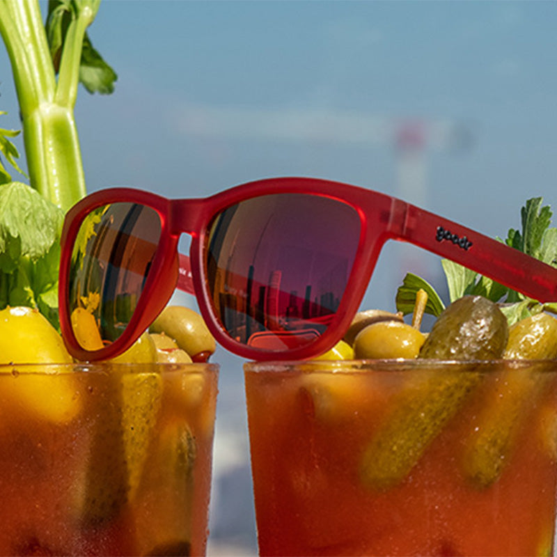Dreiviertelansicht einer roten Sonnenbrille mit roten reflektierenden Gläsern, die auf zwei Bloody-Mary-Cocktails sitzt.