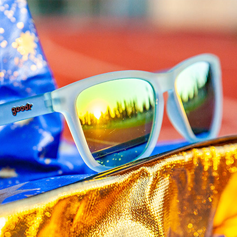 Driekwart hoekopname van een doorschijnende lichtblauwe zonnebril met goudspiegelende glazen bovenop een tovenaarshoed op een racebaan.