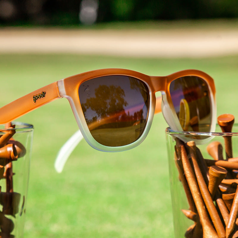 Vue de trois quarts d'angle de lunettes de soleil dégradées de brun à blanc posées sur un champ de verres à boire avec des tees de golf bruns.