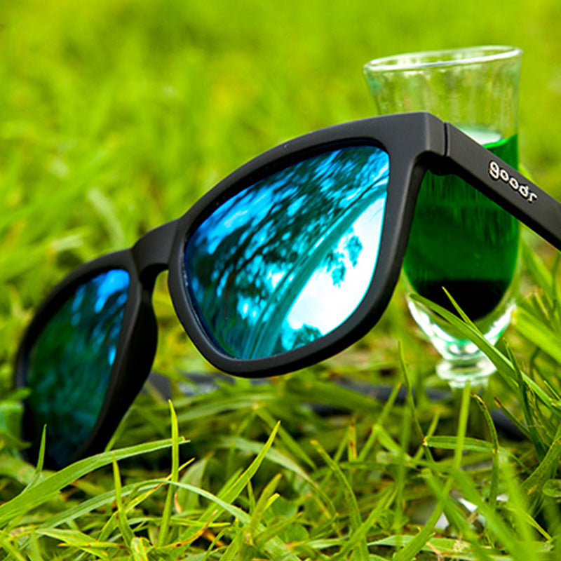 Vue de trois quarts d'angle de lunettes de soleil noires avec des verres verts réfléchissants dans un champ, un verre d'absinthe derrière elles.