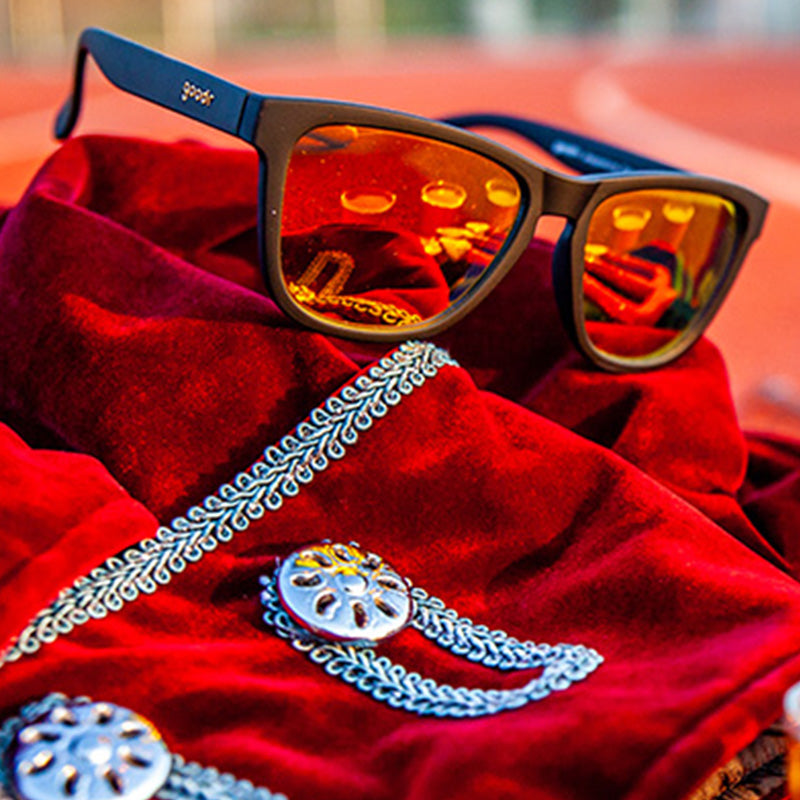 Dreiviertelansicht einer schwarzen Sonnenbrille mit bernsteinfarbenen, reflektierenden Gläsern, die auf einem roten Samtmantel auf einer Laufbahn sitzt.