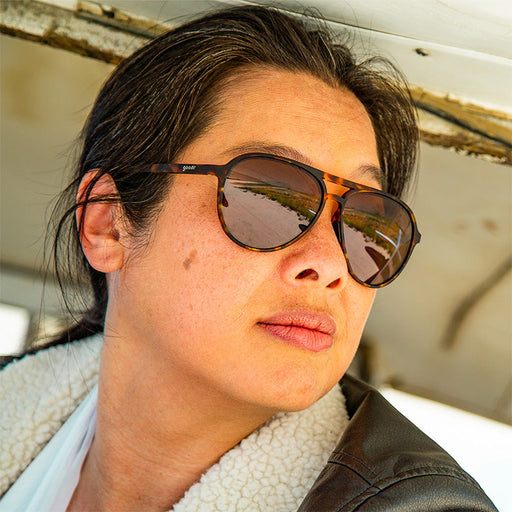 Een vrouw in een bomberjack en een bruine zonnebril met schildpadmotief kijkt naar buiten, een vliegtuighangar weerspiegeld in de glazen.
