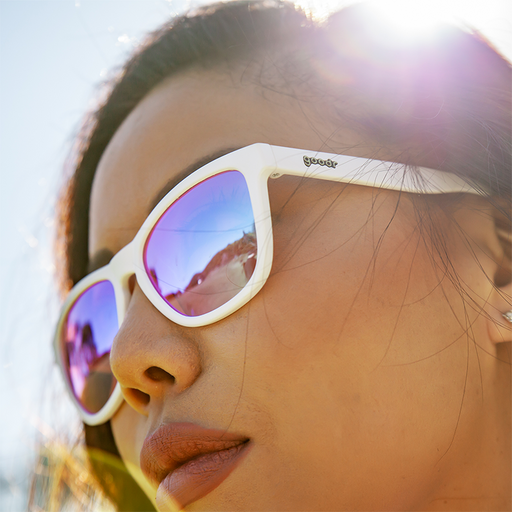 Eine Frau, die eine weiße Sonnenbrille mit nicht verspiegelten, rosafarbenen Gläsern trägt, blickt an einem sonnigen Tag grimmig zur Seite.