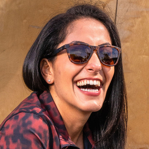 Eine Frau mit dunklem Haar, die eine braune Schildpatt-Sonnenbrille mit braunen Gläsern trägt, lacht in der Sonne.