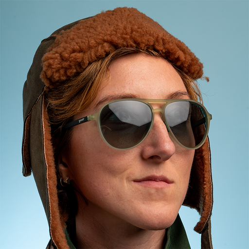 Una mujer con una gorra de aviador de pelo marrón se asoma con confianza, llevando unas aviadoras verde cadete con lentes verdes degradadas.