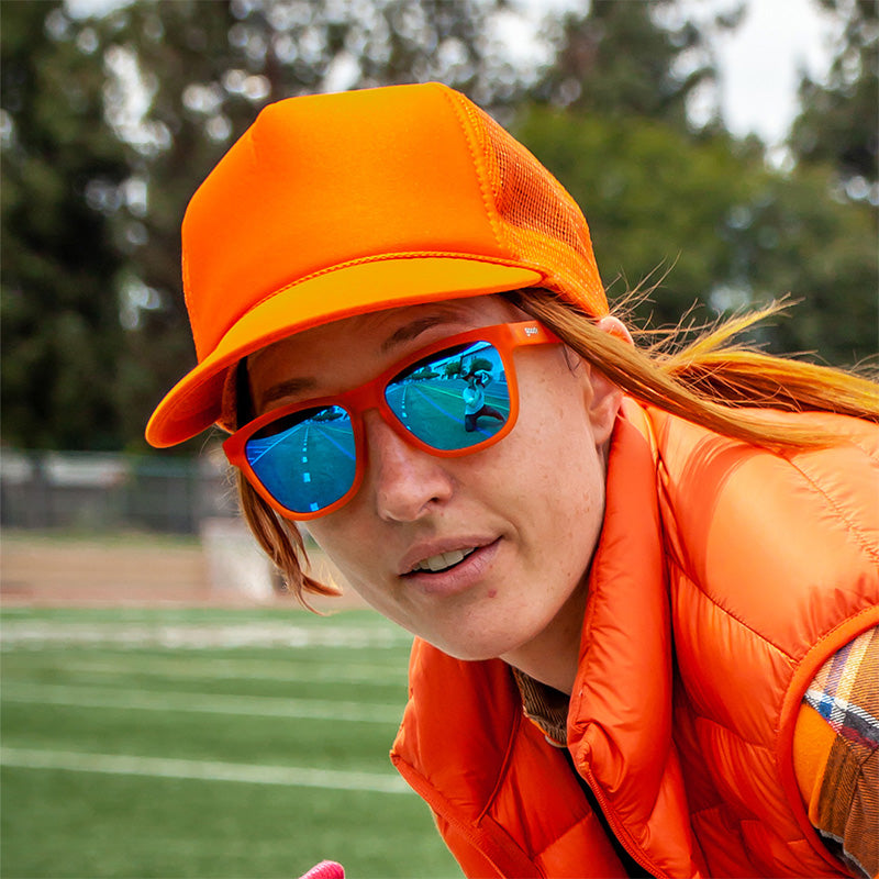 Eine Frau in einem leuchtend orangefarbenen Outfit steht auf einem Fußballfeld und trägt eine leuchtend orangefarbene Sonnenbrille mit blau verspiegelten Gläsern.