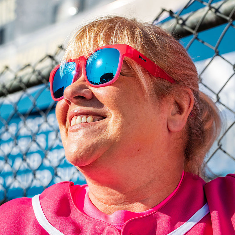 A woman in a pink baseball uniform and pink sunglasses with blue reflective lenses smiles, leaning against a fence.