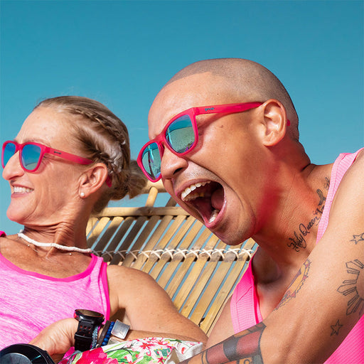 Un homme et une femme portant des lunettes de soleil rose vif avec des verres verts rient en se prélassant sur une plage tropicale.