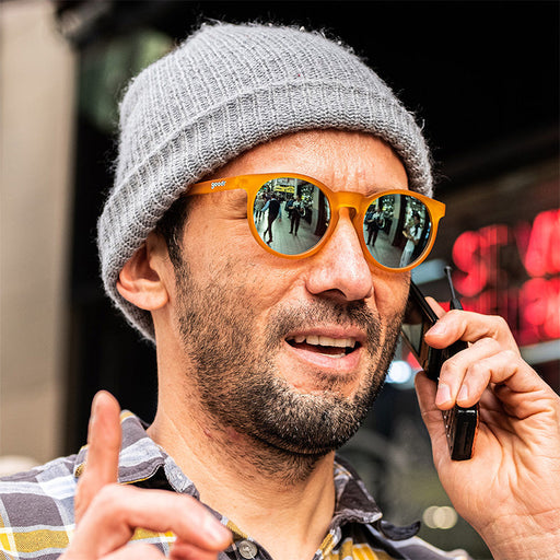 Een hippe man met een grijze muts en een gepolariseerde ronde oranje zonnebril kijkt in de verte terwijl hij praat op een fliptelefoon.