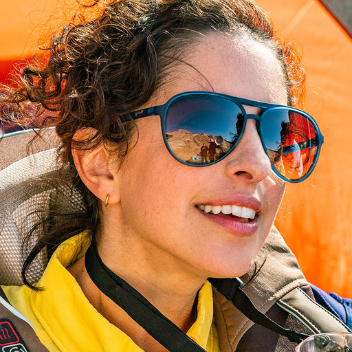 Een vrouw draagt een marineblauwe pilotenzonnebril met donkere amberkleurige glazen en glimlacht zachtjes terwijl ze in de verte kijkt.