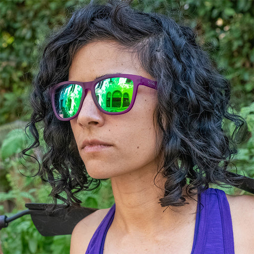 Una mujer en un jardín mira ferozmente con unas gafas de sol moradas con cristales reflectantes verdes, sosteniendo una paleta de jardín morada.