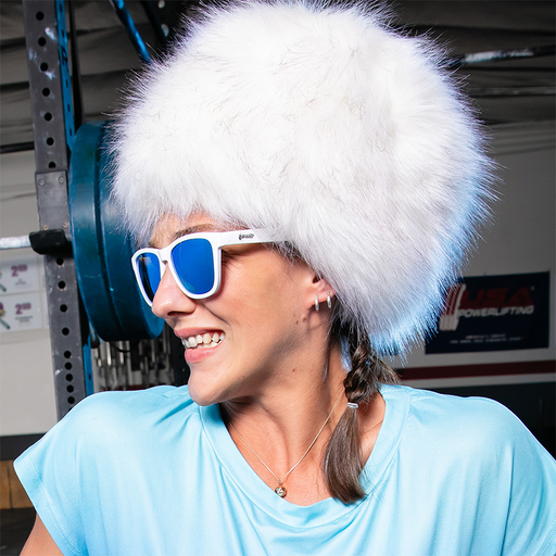 Een vrouw met een witte bontmuts en een witte zonnebril met blauwe spiegelende glazen kijkt opzij, een gewichtenbank achter haar.