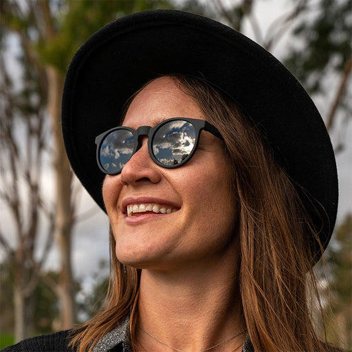 Een glimlachende hipster met een zwarte hoed en een ronde zwarte zonnebril met niet-reflecterende zwarte glazen kijkt opzij.