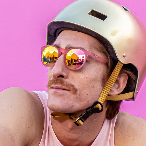 Un homme élégant, coiffé d'un casque de vélo doré, porte des lunettes de soleil rondes aux verres roses, en regardant sur le côté.