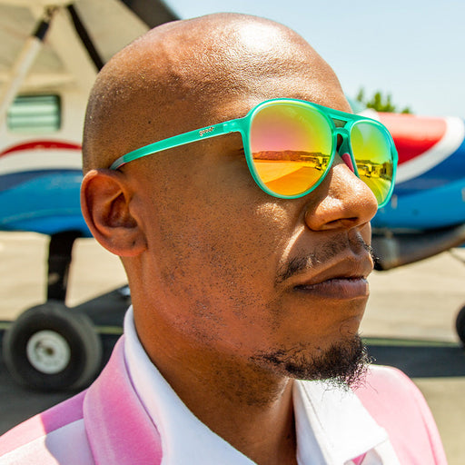 Un uomo con occhiali da sole da aviatore verde acqua con lenti rosa riflettenti guarda in lontananza, un piccolo aereo alle sue spalle.