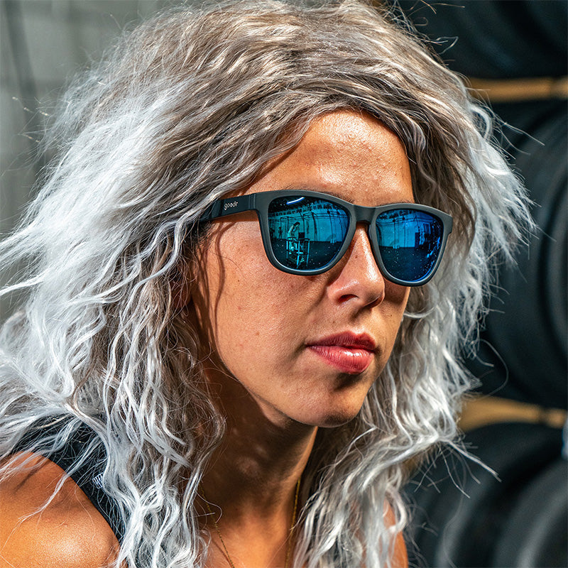 Eine Frau mit einer lockigen grauen Vokuhila-Perücke schaut zur Seite und trägt eine schwarze Sonnenbrille mit blauen, reflektierenden Gläsern.
