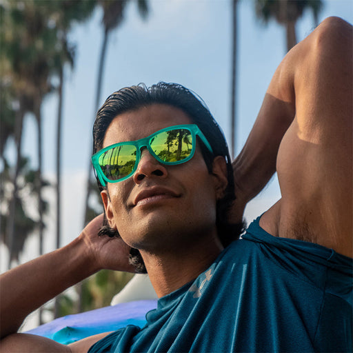 Un hombre musculoso y sonriente lleva gafas de sol con cristales reflectantes de color verde azulado y flexiona sus bíceps, con palmeras en la distancia.
